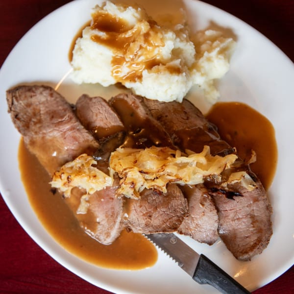 Sliced roast beef with gravy and caramelized onions, served with mashed potatoes on a white plate.