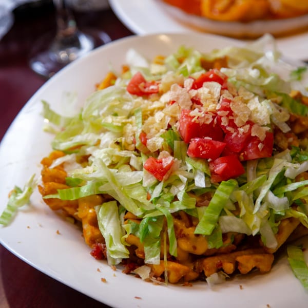 A plate of waffle fries topped with melted cheese, shredded lettuce, diced tomatoes, and crumbled chips.
