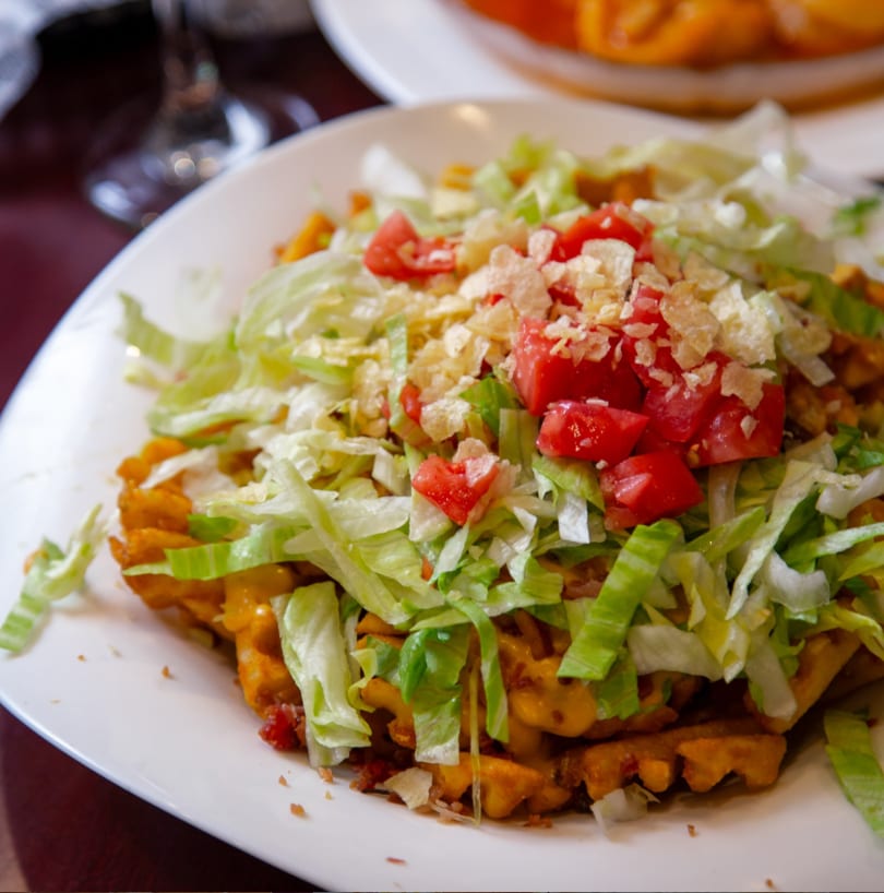 A plate of waffle fries topped with melted cheese, shredded lettuce, diced tomatoes, and crumbled chips.