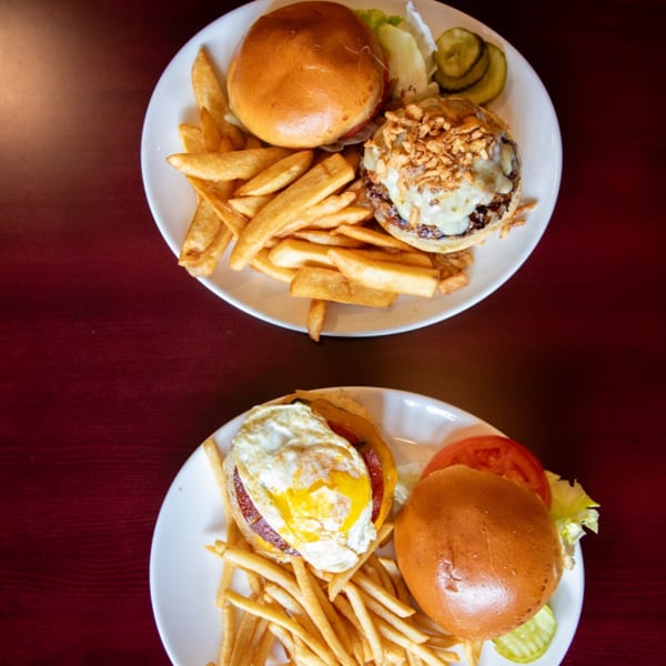 Two plates of burgers and fries, one with a fried egg, the other with crispy onions.
