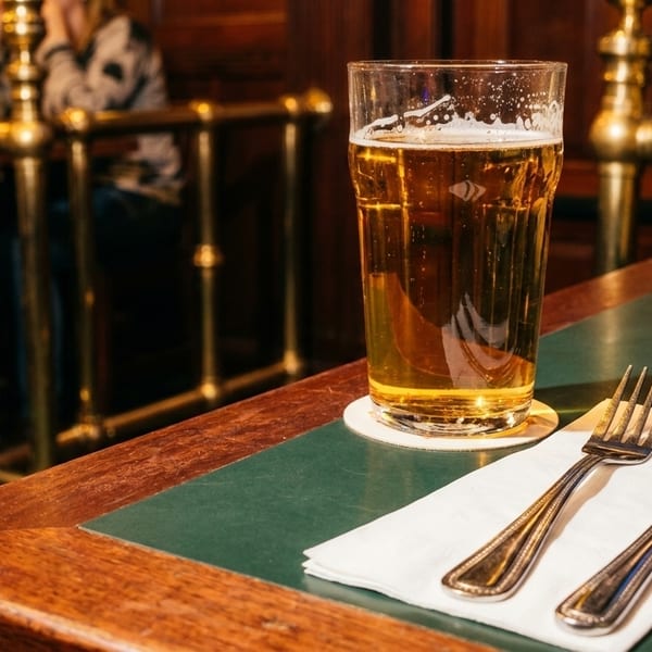 A pint of beer and cutlery on a green-topped wooden table in a dimly lit pub.