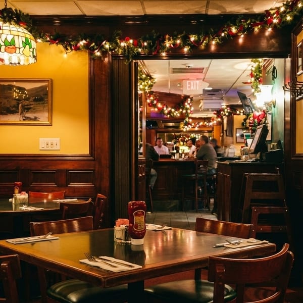 A cozy restaurant dining room with festive garland, wooden tables, and a view into a lively bar area.